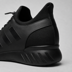 A close-up studio shot of the rear and side of a black low-top sneaker, showing matte and textured synthetic fabric panels, black laces, and a chunky black sole with diagonal ridges, set against a light gray background.