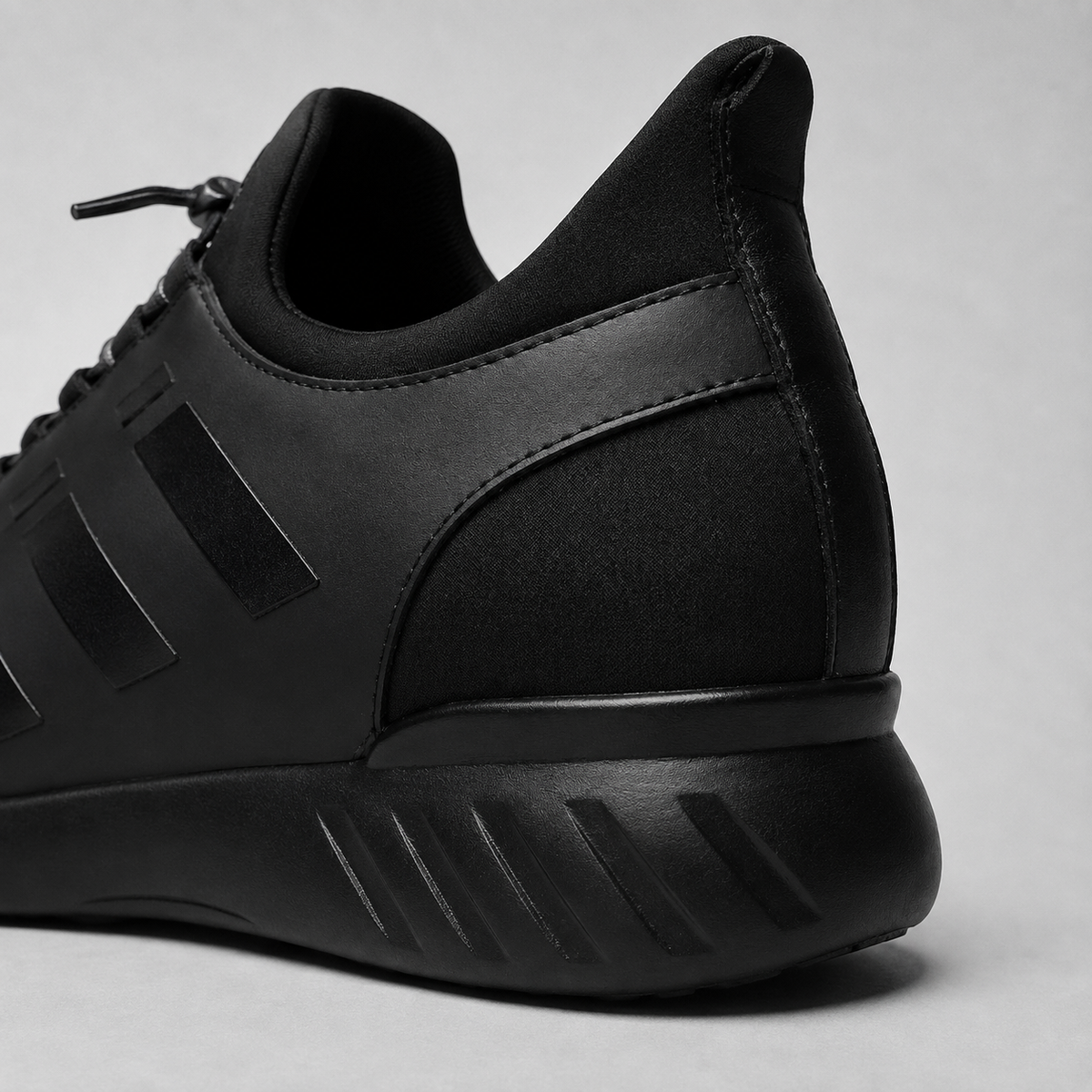 A close-up studio shot of the rear and side of a black low-top sneaker, showing matte and textured synthetic fabric panels, black laces, and a chunky black sole with diagonal ridges, set against a light gray background.