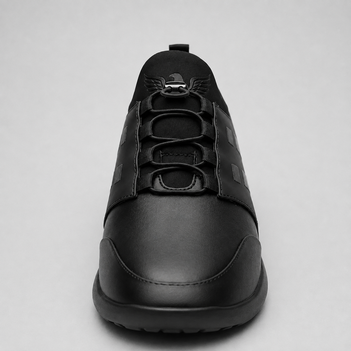 A black low-top sneaker shown from a front-facing perspective against a light gray background. The shoe features a matte black leather upper with a textured synthetic panel on the side, matching black laces, and a small winged emblem on the tongue.