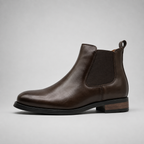 A dark brown leather Chelsea boot with a black elastic side panel and a low stacked heel, shown from a side profile on a light gray background.